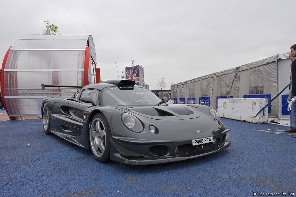 The same car parked on the blue paddock carpet at a race meeting, hospitality marquees and Michelin boards behind it.